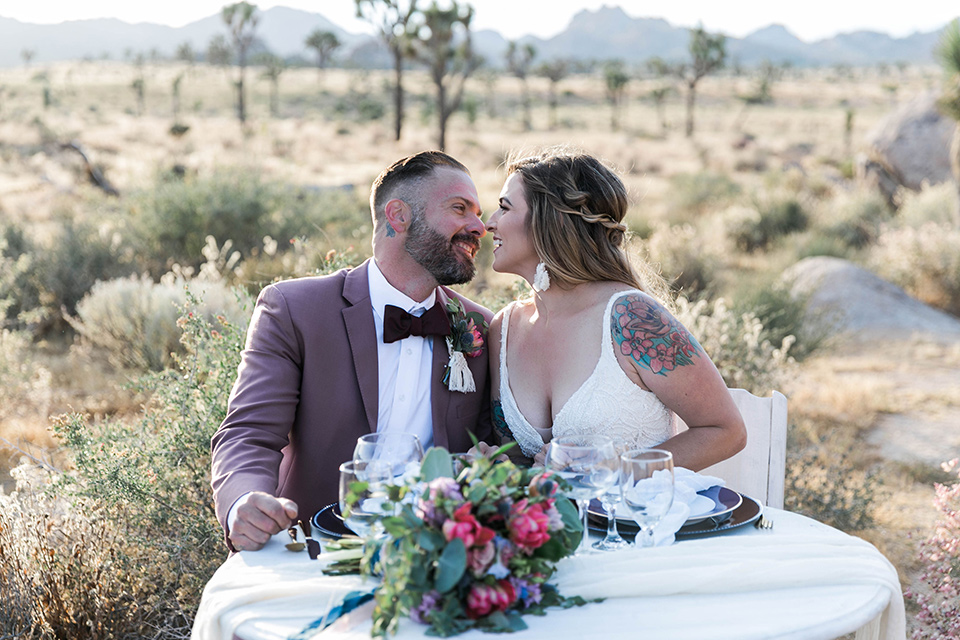  bride in a white lace bohemian style gown with a sweetheart neckline and thin straps and the groom in a rose pink suit with a burgundy bow tie sitting at sweetheart table