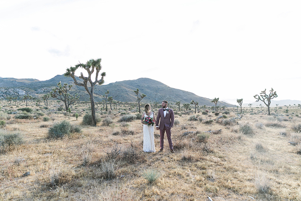  bride in a white lace bohemian style gown with a sweetheart neckline and thin straps and the groom in a rose pink suit with a burgundy bow tie walking together