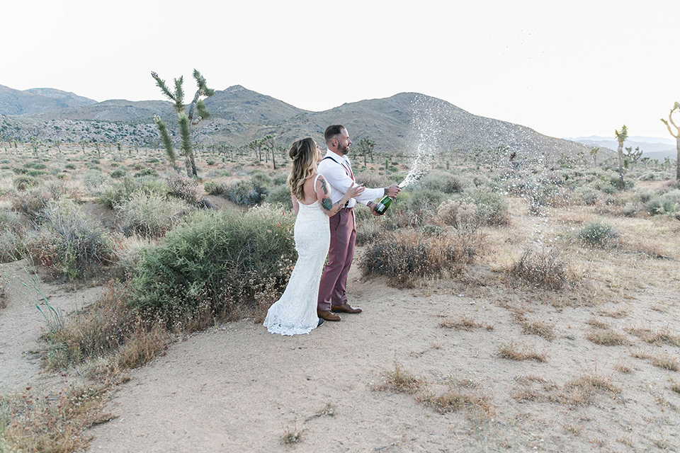  bride in a white lace bohemian style gown with a sweetheart neckline and thin straps and the groom in a rose pink suit with a burgundy bow tie walking together