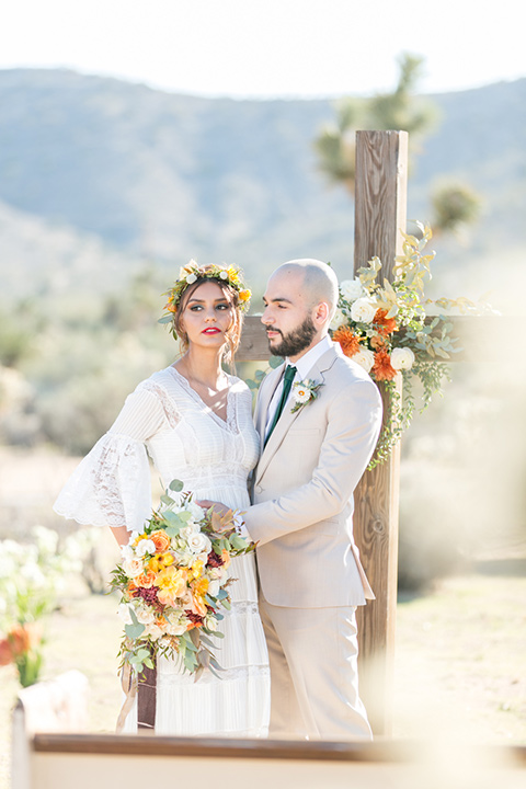  bride in a bohemian style gown with lace and a floral crown, the groom in a tan suit with a teal long tie 