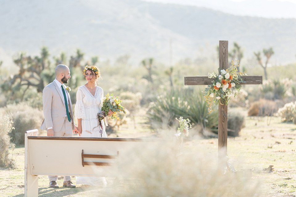  bride in a bohemian style gown with lace and a floral crown, the groom in a tan suit with a teal long tie walking