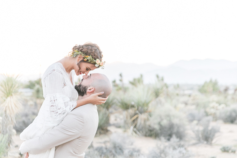  bride in a bohemian style gown with lace and a floral crown, the groom in a tan suit with a teal long tie groom lifting the bride up