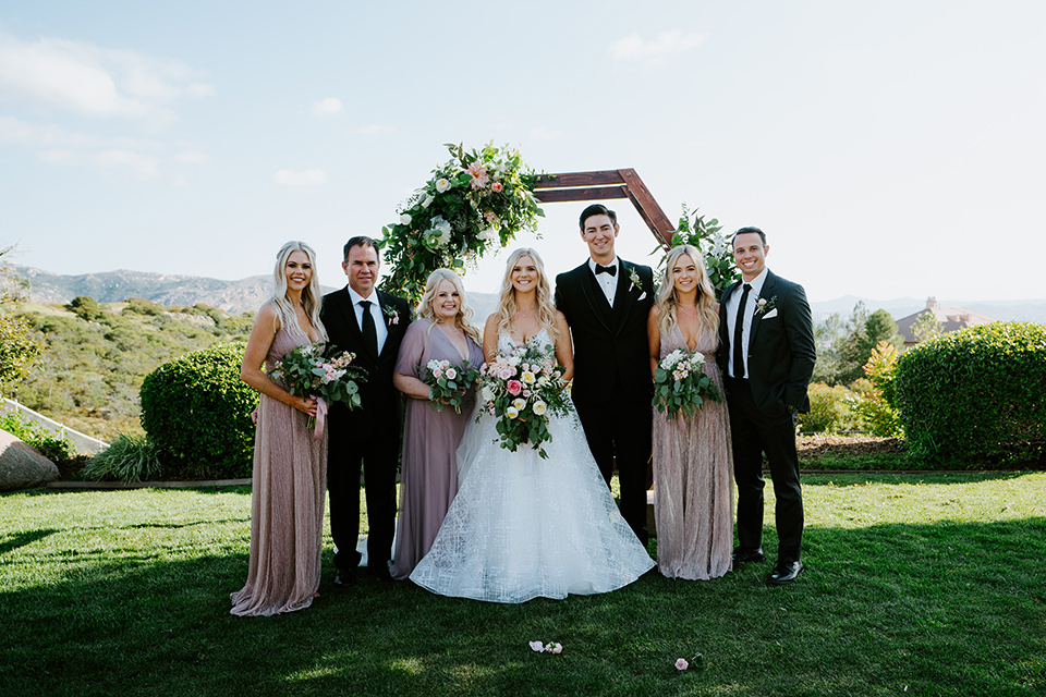  bride in a white ballgown with a strapless neckline and a long veil and the groom in a notch lapel tuxedo and black bow tie, the bridesmaids in rose gold long gowns and the groomsmen in black tuxedos with black long ties