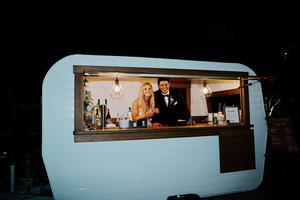  bride in a white ballgown with a strapless neckline and a long veil and the groom in a notch lapel tuxedo and black bow tie, in a trailer mobile bar