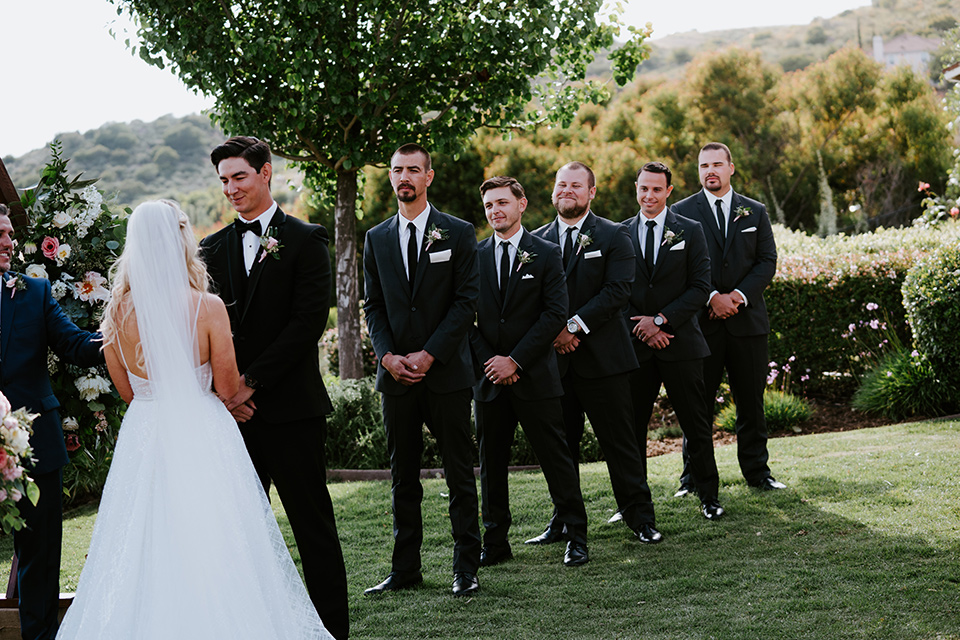  bride in a white ballgown with a strapless neckline and a long veil and the groom in a notch lapel tuxedo and black bow tie, the groomsmen in black tuxedos with black long ties at the ceremony