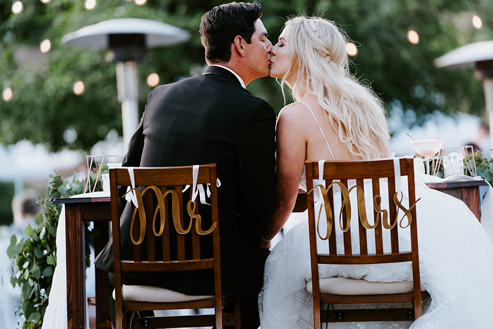  bride in a white ballgown with a strapless neckline and a long veil and the groom in a notch lapel tuxedo and black bow tie, at the reception in their chairs