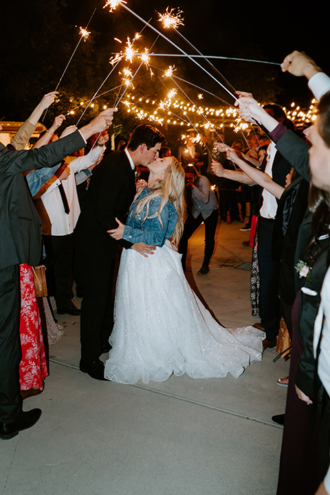 bride in a white ballgown with a strapless neckline and a long veil and jean jacket, and the groom in a notch lapel tuxedo and black bow tie kissing with the sparklers