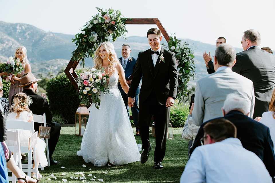  bride in a white ballgown with a strapless neckline and a long veil and the groom in a notch lapel tuxedo and black bow tie, walking down the aisle