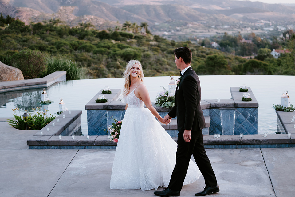  bride in a white ballgown with a strapless neckline and a long veil and the groom in a notch lapel tuxedo and black bow tie, walking by the pool