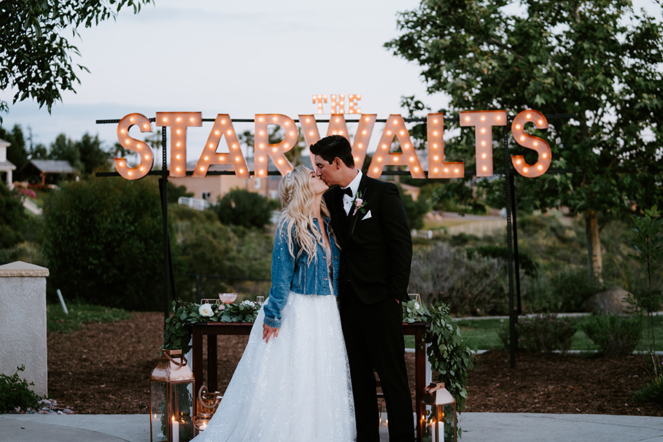  bride in a white ballgown with a strapless neckline and a long veil and a jean jacket, and the groom in a notch lapel tuxedo and black bow tie, kissing next to marquee lights