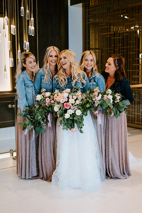 bride in a white ballgown with a strapless neckline and a long veil and jean jacket, and her bridesmaids in rose gold gowns and jean jackets