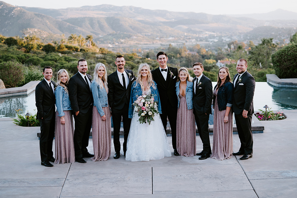  bride in a white ballgown with a strapless neckline and a long veil and jean jacket, the groom in a notch lapel tuxedo and black bow tie, the bridesmaids in rose gold long gowns and jean jackets, and the groomsmen in black tuxedos with black long ties