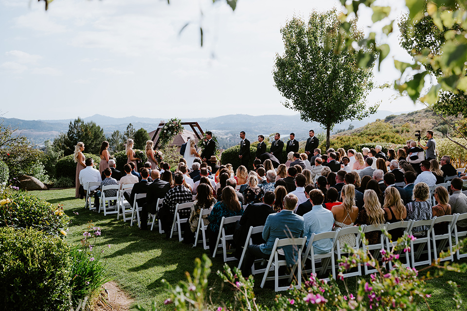  bride in a white ballgown with a strapless neckline and a long veil and jean jacket, the groom in a notch lapel tuxedo and black bow tie, the bridesmaids in rose gold long gowns and jean jackets, and the groomsmen in black tuxedos with black long ties at the ceremony