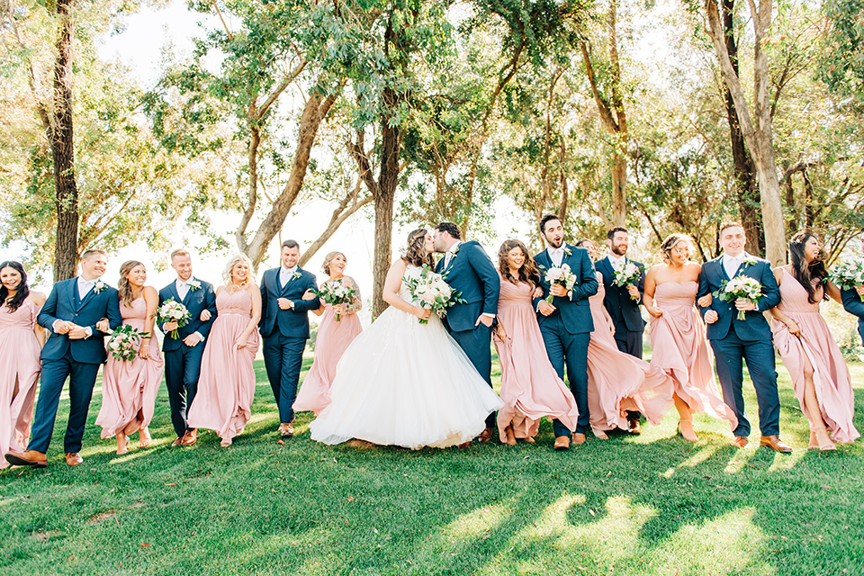  bride in a white ball gown with train and the groom in a dark blue suit in bow tie, bridesmaids in blush gowns and the groomsmen in dark blue suits