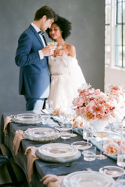  bride in a tulle ballgown with an off the shoulder detail and full billowed sleeves and the groom in a slate blue suit with a matching bow tie at the end of the table