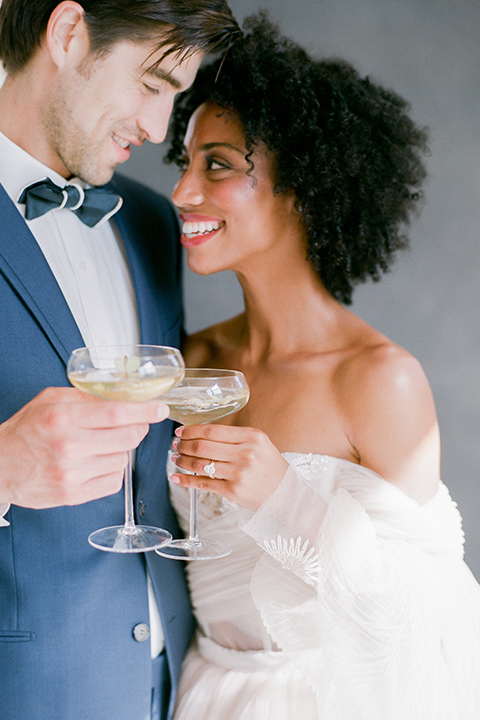  bride in a tulle ballgown with an off the shoulder detail and full billowed sleeves holding cocktail glasses