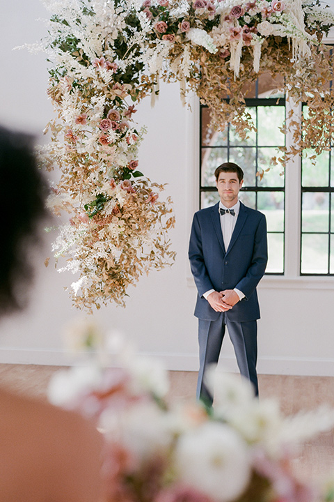  bride in a tulle ballgown with an off the shoulder detail and full billowed sleeves and the groom in a slate blue suit with a matching bow tie waiting at the end of the altar for bride