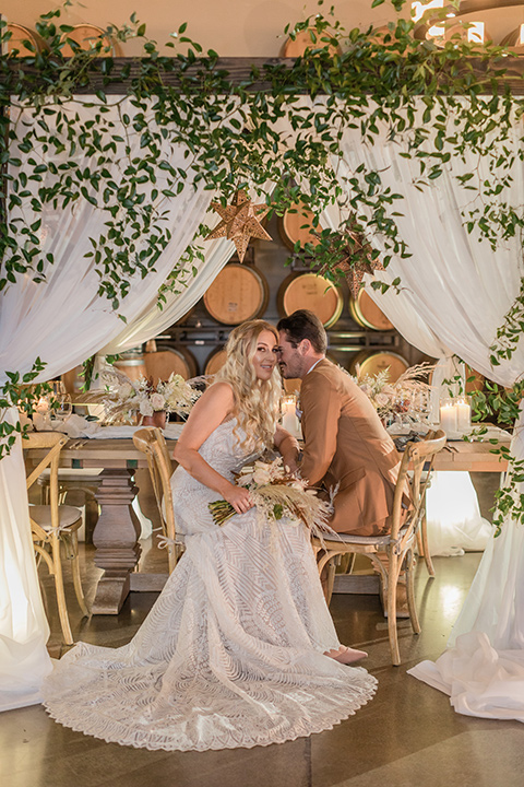   bride in a lace bohemian gown and the groom in a caramel brown suit with a chocolate brown long tie and fun pocket square sitting at the reception table 