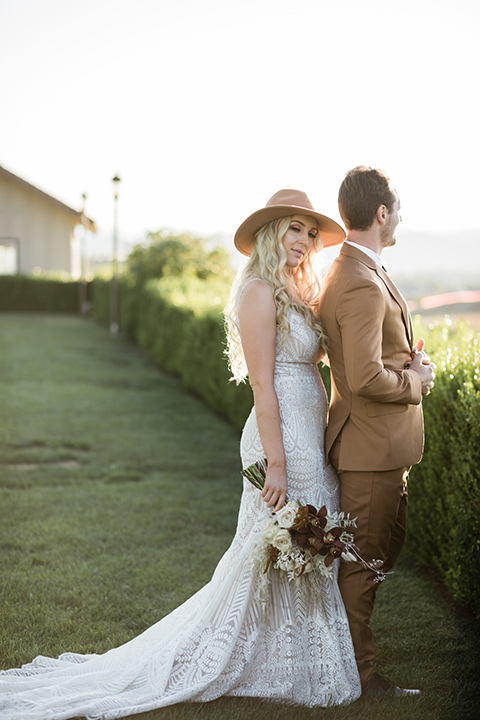   bride in a lace bohemian gown and the groom in a rust colored suit with a deep brown long tie and fun pocket square standing outside venue 
