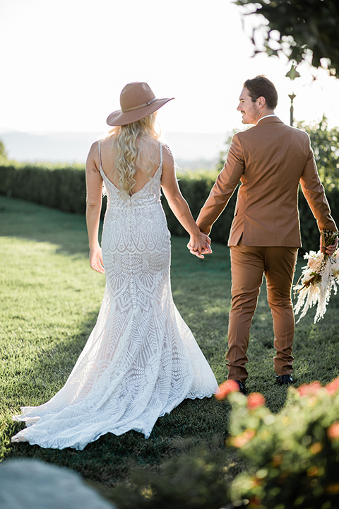  bride in a lace bohemian gown and wide brimmed hat and the groom in a burnt orange suit with a brown long tie and fun pocket square together outside at the winery 