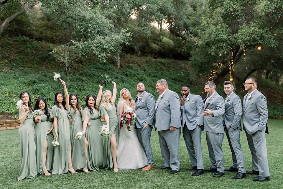  bride in a mermaid style lace white gown and the groom in a light grey suit with a light brown shoes and white pocket square, the bridesmaids in sage green dresses and the groomsmen in light grey suits and bow ties