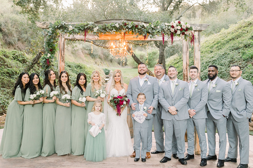  bride in a mermaid style lace white gown and the groom in a light grey suit with a light brown shoes and white pocket square, the bridesmaids in sage green dresses and the groomsmen in light grey suits and bow ties