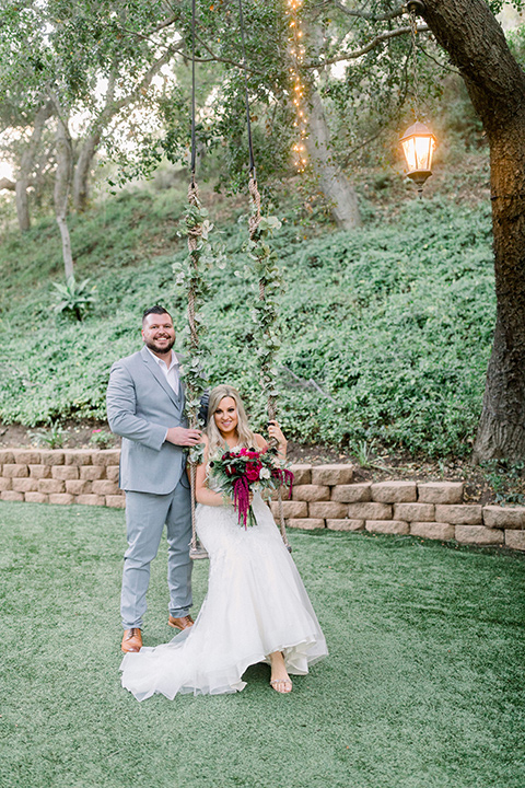  bride in a white lace mermaid style gown and a strapless neckline and the groom in a light grey suit on a tree swing