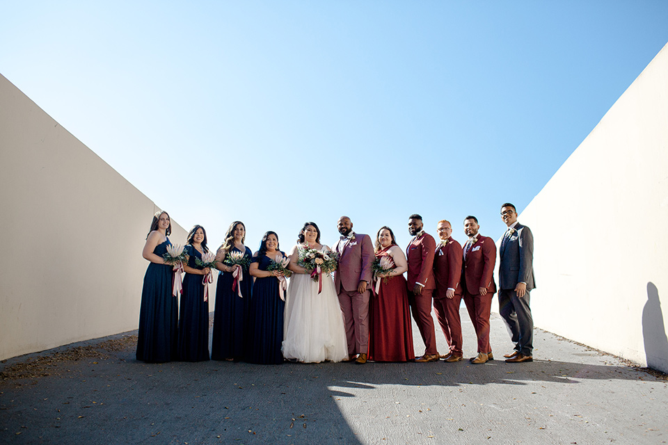  bride in a tulle ballgown with cap sleeves and the groom in a rose pink suit, the groomsmen in burgundy suits and the bridesmaids in black gowns