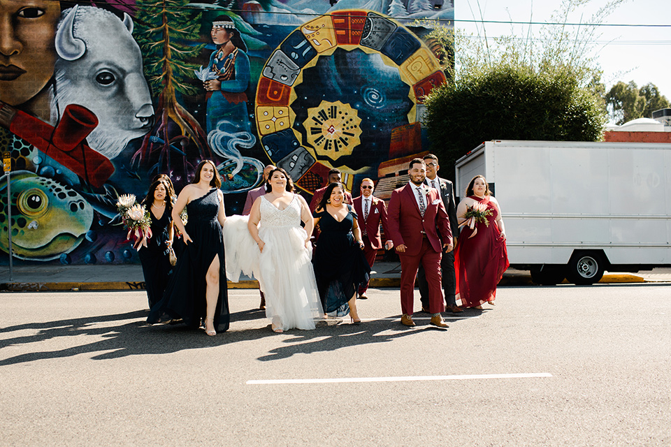  bride in a tulle ballgown with cap sleeves and the groom in a rose pink suit, the groomsmen in burgundy suits and the bridesmaids in black gowns, walking across the street