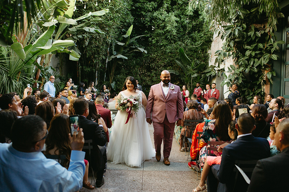  bride in a tulle ballgown with cap sleeves and the groom in a rose pink suit walking down the aisle at the ceremony