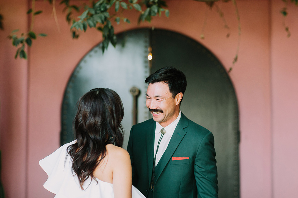  bride in a white bohemian gown with one sleeve and a ruffled detail with a pink veil and the groom in a green suit with a white shirt, bolo tie, and wide brimmed hat 