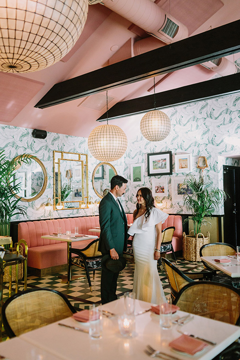  bride in a white bohemian gown with one sleeve and a ruffled detail with a pink veil and the groom in a green suit with a white shirt, bolo tie, and wide brimmed hat in an eccentric pink and mustard colored room with fun paintings on the wall