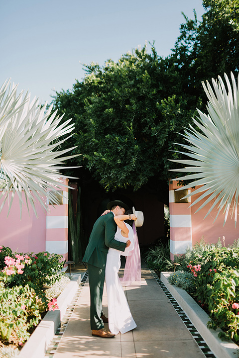  bride in a white bohemian gown with one sleeve and a ruffled detail the groom in a green suit with a white shirt and bolo tie kissing outside venue