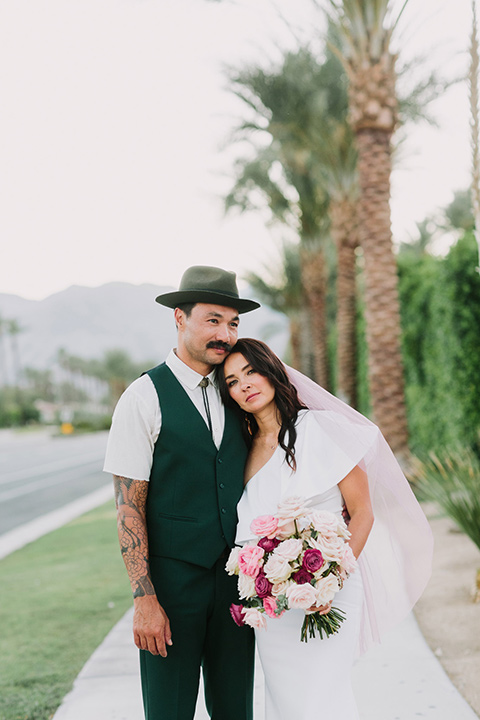  bride in a white bohemian gown with one sleeve and a ruffled detail with a pink veil and the groom in a green suit with a white shirt, bolo tie, and wide brimmed hat