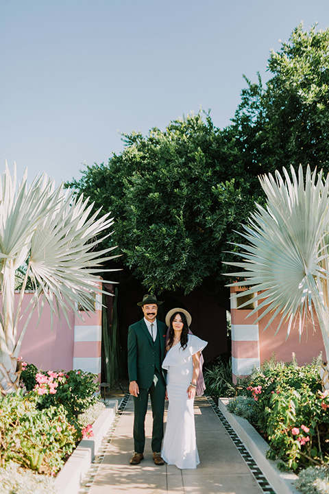  bride in a white bohemian gown with one sleeve and a ruffled detail with a pink veil and the groom in a green suit with a white shirt, bolo tie, and wide brimmed hat looking at camera outside venue