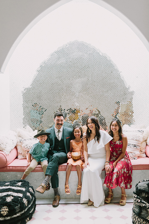  bride in a white bohemian gown with one sleeve and a ruffled detail the groom in a green suit with a white shirt and bolo tie sitting inside with their children around them