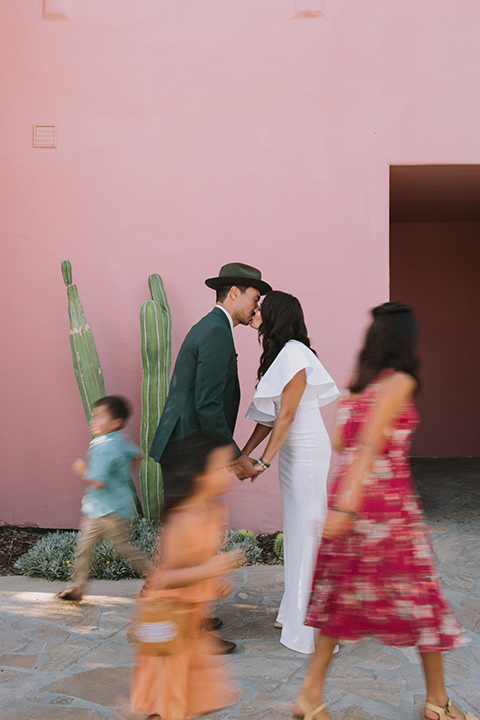  bride in a white bohemian gown with one sleeve and a ruffled detail with a pink veil and the groom in a green suit with a white shirt, bolo tie, and wide brimmed hat kissing with their children running around them