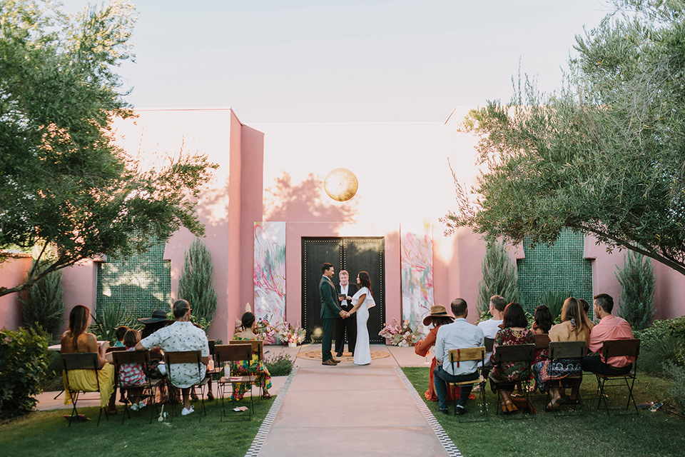  bride in a white bohemian gown with one sleeve and a ruffled detail with a pink veil and the groom in a green suit with a white shirt, bolo tie, and wide brimmed hat at the ceremony 