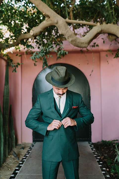  the groom in a green suit with a white shirt and bolo tie and wide brimmed hat buttoning his jacket