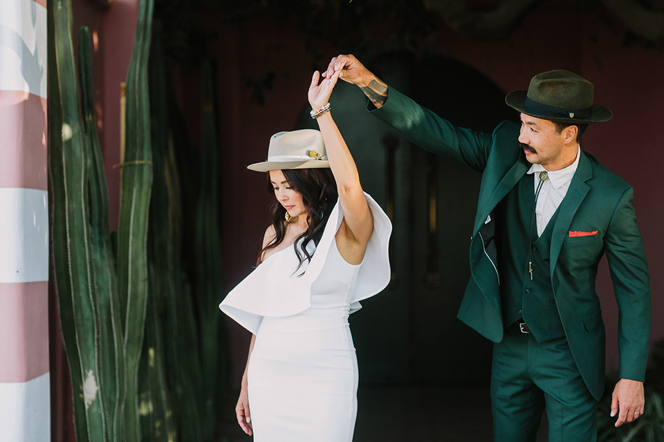  bride in a white bohemian gown with one sleeve and a ruffled detail with a pink veil and the groom in a green suit with a white shirt, bolo tie, and wide brimmed hat dancing 