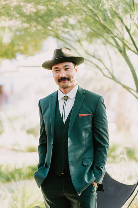  the groom in a green suit with a white shirt, bolo tie, and wide brimmed hat smiling at the camera