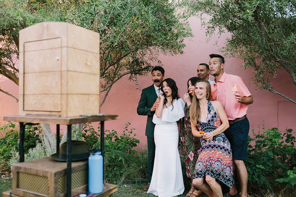  bride in a white bohemian gown with one sleeve and a ruffled detail with a pink veil and the groom in a green suit with a white shirt, bolo tie, and wide brimmed hat dancing smiling at a photobooth 