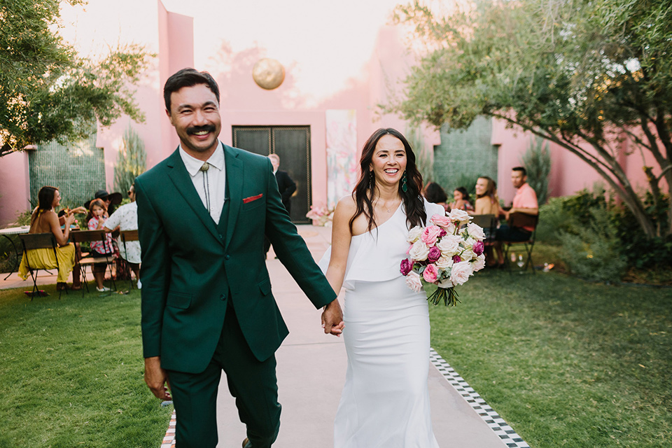  bride in a white bohemian gown with one sleeve and a ruffled detail with a pink veil and the groom in a green suit with a white shirt, bolo tie, and wide brimmed hat walking down the aisle