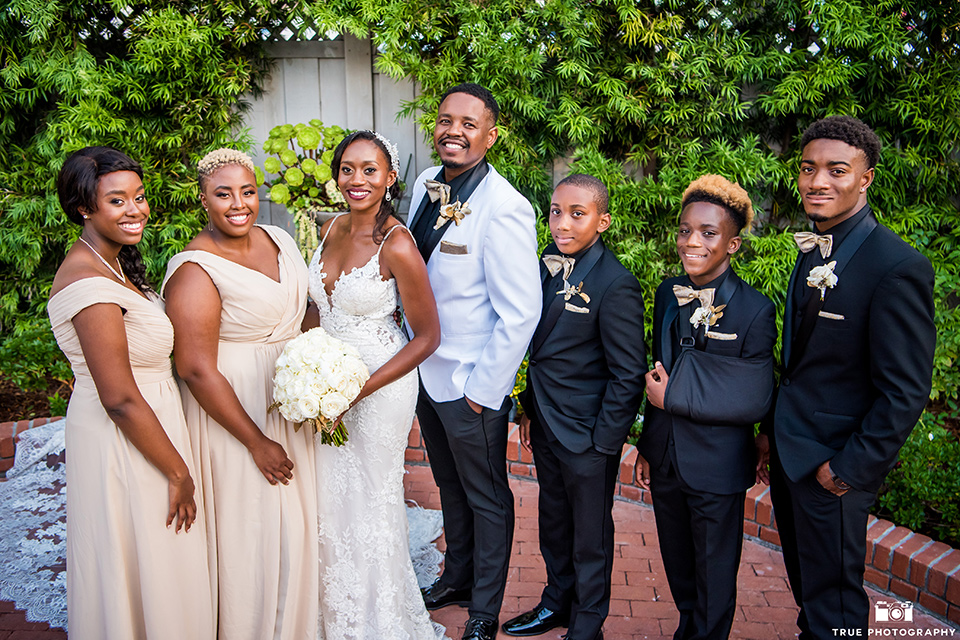  bride in a white formfitting gown with a plunging neckline and lace detailing and the groom in a white shawl lapel tuxedo with a black shirt and gold bow tie, the bridesmaids in a gold long gowns and the groomsmen in all black tuxedos and gold bow ties