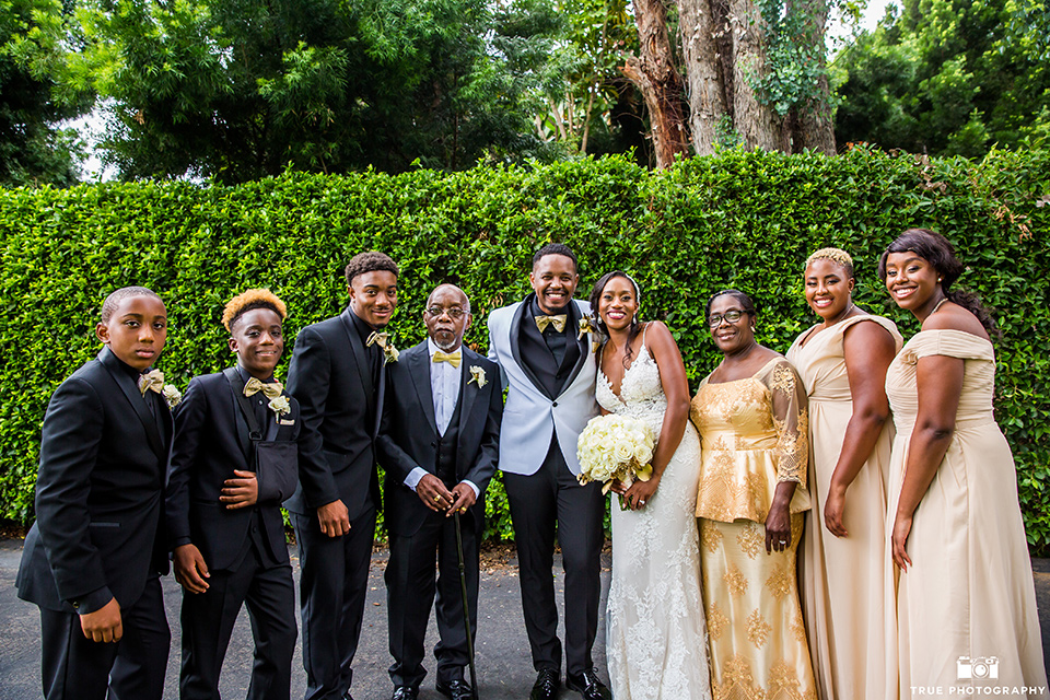  bride in a white formfitting gown with a plunging neckline and lace detailing and the groom in a white shawl lapel tuxedo with a black shirt and gold bow tie, the bridesmaids in a gold long gowns and the groomsmen in all black tuxedos and gold bow ties