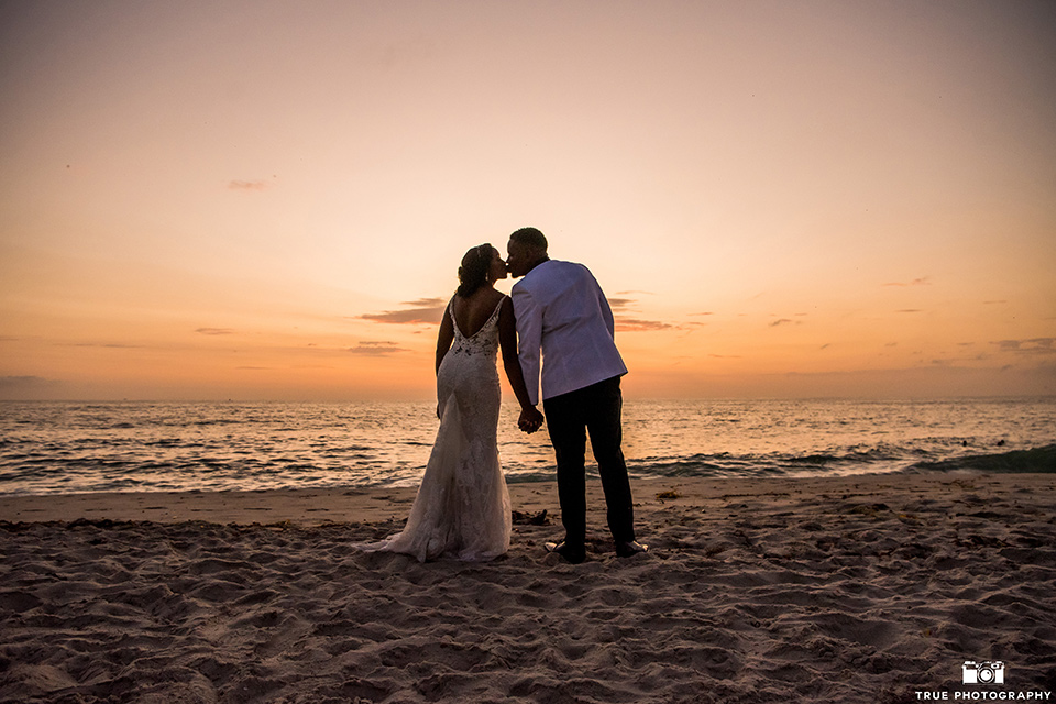  bride in a white formfitting gown with a plunging neckline and lace detailing and the groom in a white shawl lapel tuxedo with a black shirt and gold bow tie, at sunset