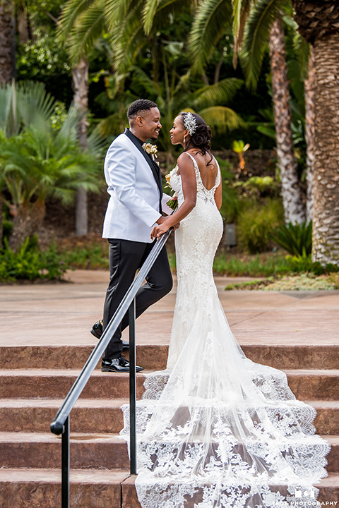  bride in a white formfitting gown with a plunging neckline and lace detailing and the groom in a white lace tuxedo with a black shirt and gold bow tie, kissing on the steps