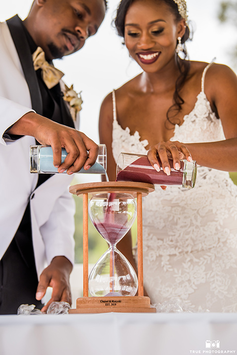  bride in a white formfitting gown with a plunging neckline and lace detailing and the groom in a white lace tuxedo with a black shirt and gold bow tie, with sand at ceremony