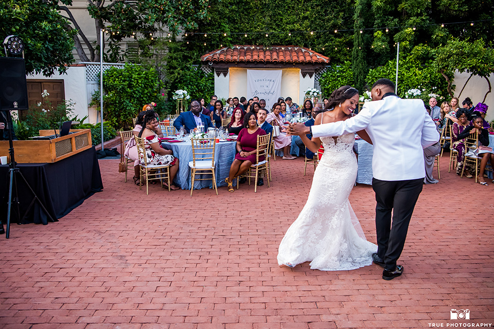  bride in a white formfitting gown with a plunging neckline and lace detailing and the groom in a white shawl lapel tuxedo with a black shirt and gold bow tie, first dance