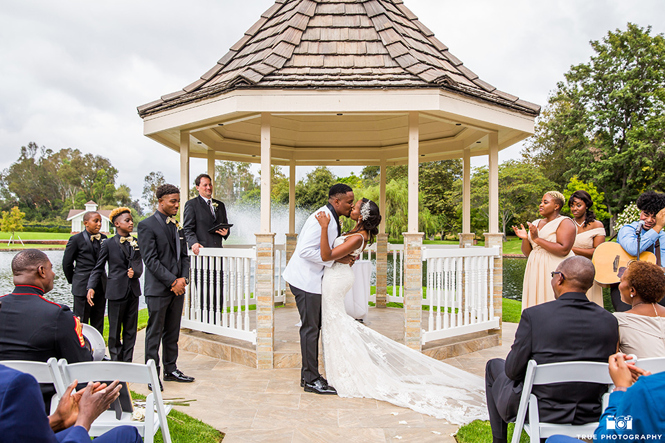  bride in a white formfitting gown with a plunging neckline and lace detailing and the groom in a white shawl lapel tuxedo with a black shirt and gold bow tie, first kiss